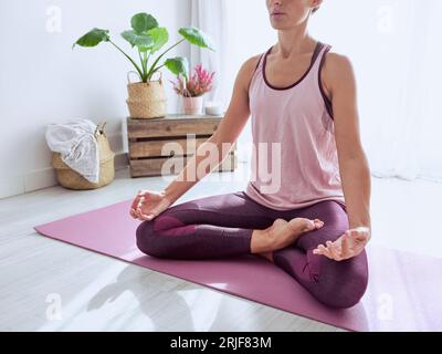 Donna irriconoscibile in abbigliamento sportivo seduta su un tappetino da yoga in posa di loto con le mani strette nel mudra di gyan in una stanza leggera la mattina Foto Stock