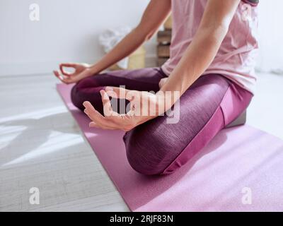 Ritaglia una donna anonima che medita nella postura di Padmasana con il gesto della mudra di Gyan durante la sessione di yoga su tappetino in una stanza luminosa a casa Foto Stock