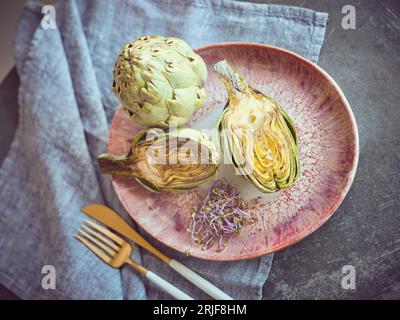 Dall'alto di deliziosi carciofi verdi con erbe servite su piatto su tavolo di marmo con posate e asciugamano in cucina Foto Stock