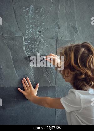 Vista posteriore di un bambino intelligente che risolve equazioni matematiche su un muro di pietra nera con un pezzo di gesso Foto Stock