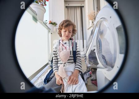 Vista dalla lavatrice di un bambino frustrato, cosparso di vestiti sporchi seduti sul pavimento in una lavanderia leggera Foto Stock