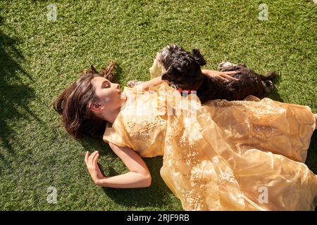 Vista dall'alto di un'affascinante ragazza con un soffice abito sdraiata sul prato verde e abbracciando l'adorabile Schnauzer in miniatura di razza pura nelle soleggiate giornate estive Foto Stock