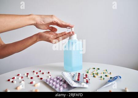 Coltura anonima persona che applica liquido antibatterico a mano vicino al tavolo con varie pillole e termometro durante la pandemia di coronavirus su sfondo bianco Foto Stock