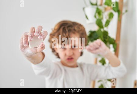 Un ragazzo dai capelli ricci sfocati che fa un volto divertente mentre mostra una barretta di sapone e un pugno stringente durante la routine di igiene quotidiana Foto Stock