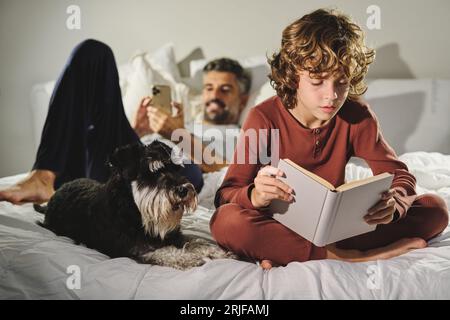 Padre con telefono sdraiato su un comodo letto vicino al figlio che legge un'interessante favola vicino al cane Schnauzer in miniatura in camera da letto a casa Foto Stock