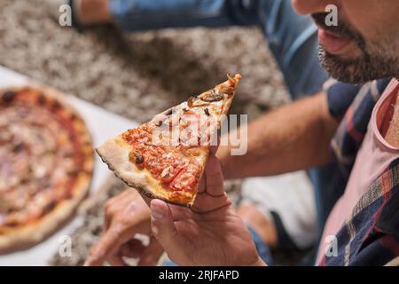 Un maschio anonimo che mangia una deliziosa pizza con fette di prosciutto alla festa Foto Stock