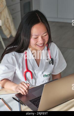 Dall'alto di allegro medico asiatico femminile in uniforme facendo consultazione online con paziente su portatile durante il lavoro in clinica Foto Stock