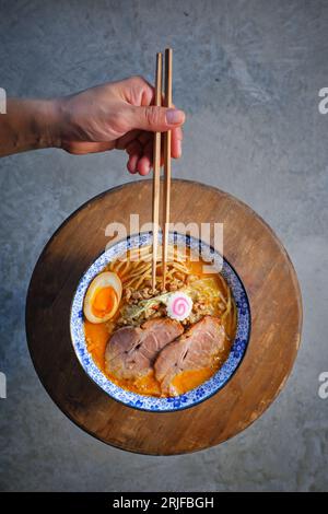 Dall'alto della persona senza volto raccolto che mangia tradizionale zuppa di noodle Asiatica con maiale Char siu e uovo servito in ciotola su tavola di legno Foto Stock