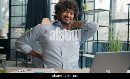 Malato uomo arabo-indiano latino-americano esausto sente dolore alla schiena dopo il lavoro del portatile male male ragazzo uomo di ufficio che massaggia doloroso collo Foto Stock