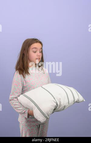 Una ragazza adolescente in pigiama si sta preparando per andare a letto. La ragazza guarda con ansia il cuscino. Foto studio, verticale, sfondo lilla. Foto Stock