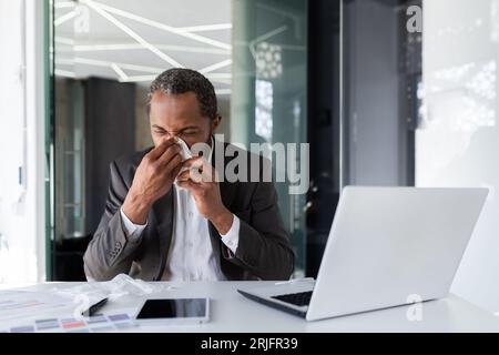 Uomo malato sul posto di lavoro all'interno dell'ufficio con gioia e freddo, uomo d'affari starnutisce seduto alla scrivania, uomo afro-americano con freddo usa il computer portatile al lavoro. Foto Stock