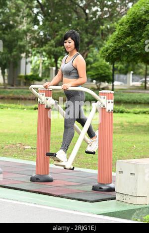 Bella donna asiatica che fa esercizio sportivo con un tipo di macchina per esercizi che oscilla le gambe nel parco all'aperto. Foto Stock