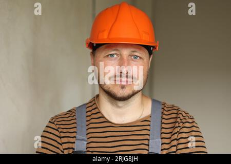 Appartamento ristrutturato. Il ritratto di un riparatore professionista barbuto e sicuro in un elmetto di sicurezza arancione e indumenti da lavoro guarda la fotocamera. Vuoto co Foto Stock