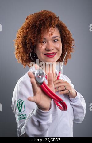 Donna veterinaria, bella con i capelli rossi, che mostra uno stetoscopio alla macchina fotografica in uniforme bianca. Isolato su sfondo grigio. Foto Stock