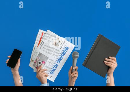 Mani femminili con telefono cellulare, giornali, microfono e notebook su sfondo blu Foto Stock