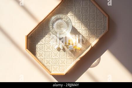 Bicchiere d'acqua e diverse capsule vitaminiche su vassoio alla luce del mattino. Foto Stock