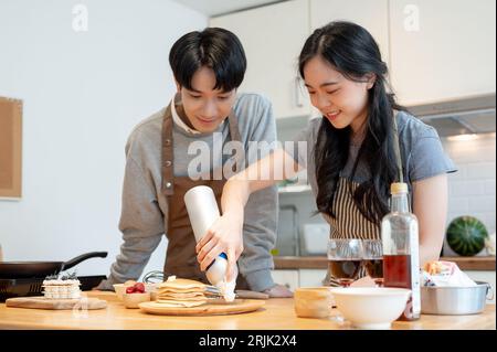 Una giovane coppia asiatica deliziosa e gioiosa prepara la colazione fatta in casa, spruzzando panna montata sui pancake e si diverte a fare i pancake nella tog della cucina Foto Stock