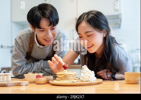 A lovely young Asian couple prepares homemade breakfast and enjoys making pancakes in the kitchen together. cooking date, boyfriend and girlfriend, ro Stock Photo