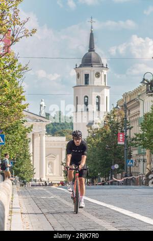 Ragazzo in bicicletta con casco sulla strada ciclabile nel centro della città di Vilnius, Lituania, con campanile e cattedrale sullo sfondo, verticale Foto Stock