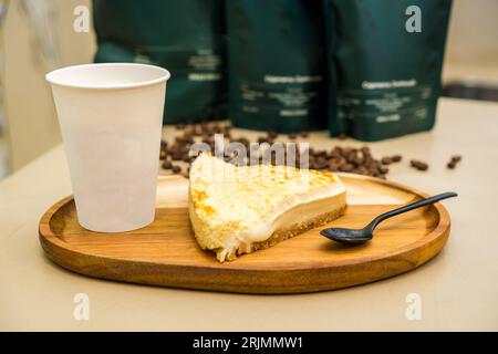 Un primo piano di una cheesecake con una tazza di caffè in plastica su un vassoio di legno, perfetto per un dolce Foto Stock