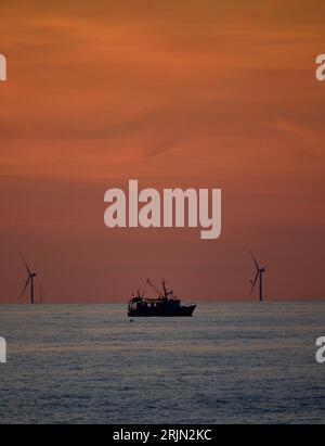 Un paesaggio marino con una nave e una linea di turbine eoliche che si stagliano contro il cielo al tramonto. Foto Stock