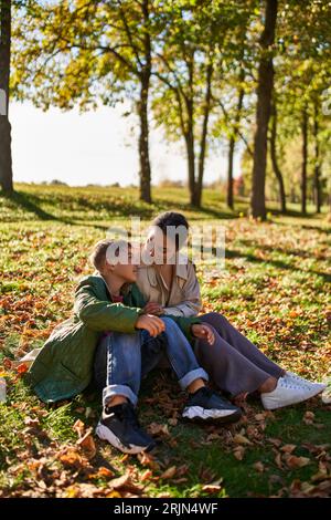 amore materno, felice donna afroamericana e figlio seduto sull'erba con foglie d'oro, autunno Foto Stock