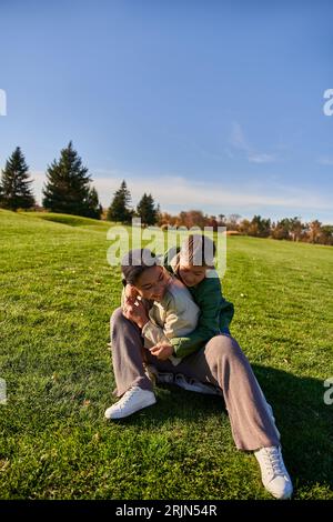madre e figlio gioiosi seduti sull'erba, giorno di sole, autunno, ragazzo afro-americano allegro che abbraccia la mamma Foto Stock