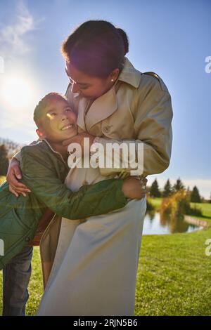 felice donna afroamericana che abbraccia figlio, in piedi vicino allo stagno nel parco, famiglia afroamericana Foto Stock