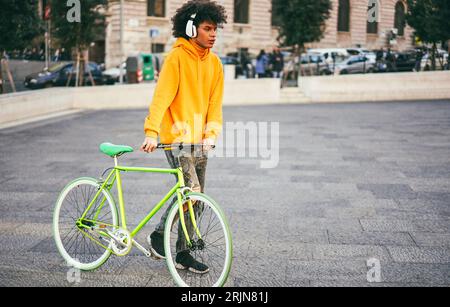 Giovane africano che gira per la città in bicicletta ascoltando musica con le cuffie - stile di vita urbano e concetto autunnale - Focus on Face Foto Stock