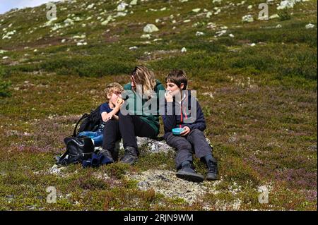 La madre con i bambini si siede su un masso e mangia frutta durante una passeggiata in montagna. Vista frontale della famiglia naturale felice con uno stile di vita sano Foto Stock