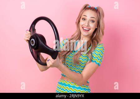 Ritratto di una bellissima ragazza impressionata con code vestite di abito colorato che regge il volante alla scuola guida isolata su sfondo rosa Foto Stock