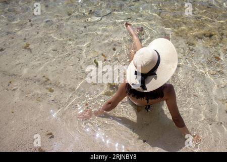 Una giovane donna con un elegante cappello e un bikini in due pezzi seduto in acque turchesi poco profonde Foto Stock