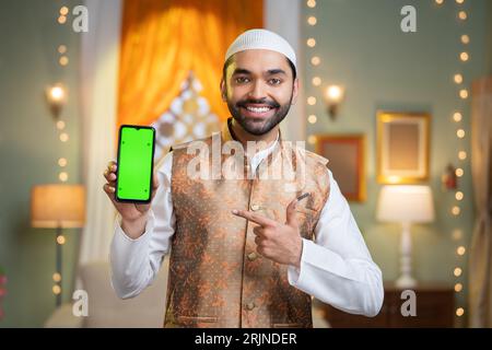 Felice giovane musulmano indiano che mostra un telefono cellulare con schermo verde puntando il dito mentre guarda la macchina fotografica - concetto di pubblicità del festival , app Foto Stock