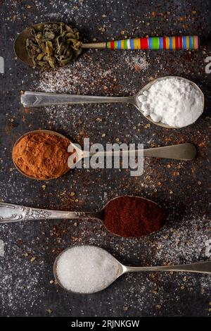 Cucchiai con tè verde, cacao, zucchero a velo e caffè macinato su fondo nero cosparso di zucchero di canna e bianco, caffè e cacao in polvere. Foto Stock