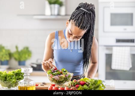 Giovane donna afroamericana prepara insalata sana, aggiunge pomodori. Foto Stock