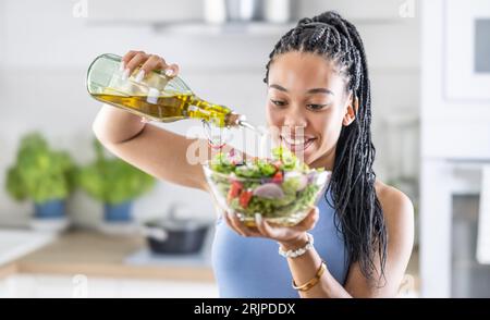 Una giovane donna afroamericana prepara un'insalata a foglia salutare, condendola con olio d'oliva. Foto Stock