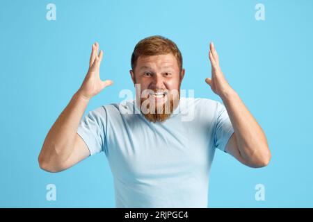 L'uomo rosso barbuto urla emotivamente, esprimendo rabbia e irritazione sullo sfondo blu dello studio Foto Stock