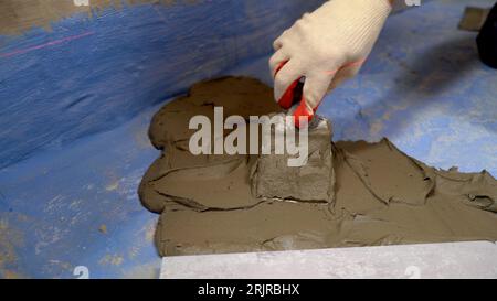 Ristrutturazione del bagno con adesivo per piastrelle. Un lavoratore poggia piastrelle per pavimenti in ceramica grigio scuro su una superficie adesiva. Adesivo per piastrelle per incollare piastrelle in ceramica a. Foto Stock