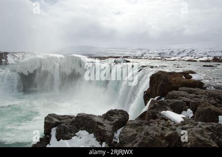 Una maestosa cascata che scende lungo il letto di un fiume roccioso in Islanda, con rocce innevate su ogni lato del fiume Foto Stock