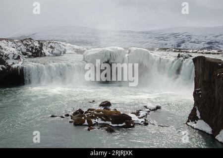 Una maestosa cascata che scende lungo il letto di un fiume roccioso in Islanda, con rocce innevate su ogni lato del fiume Foto Stock