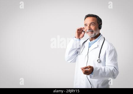 Positive caucasian senior doctor therapist in white coat and headphones consults client remotely Foto Stock