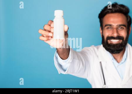Allegro medico maschio indiano che mostra barattolo di pillole alla fotocamera su sfondo blu studio, messa a fuoco selettiva, mockup Foto Stock