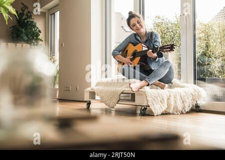 Donna sorridente seduta alla finestra a casa a suonare la chitarra Foto Stock