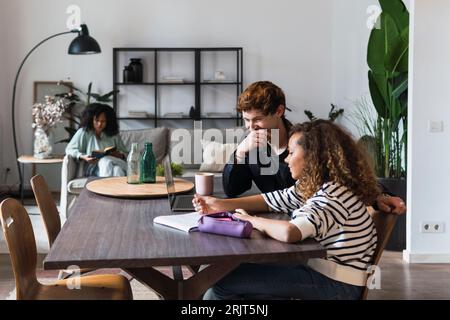 Padre che guarda la figlia fare i compiti con la madre che legge in sottofondo Foto Stock
