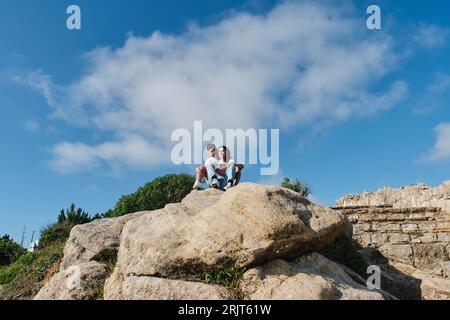 Giovane coppia seduta su rocce sotto il cielo Foto Stock