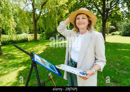 Donna matura sorridente che indossa un cappello con pitture ad acquerello nel parco Foto Stock