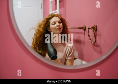 Donna che asciuga i capelli guardando allo specchio a casa Foto Stock