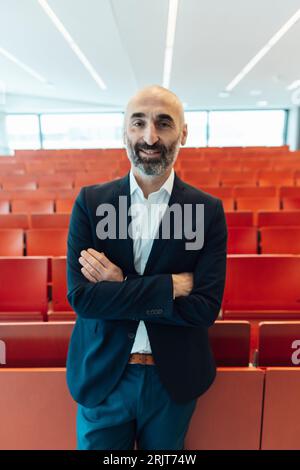 Uomo d'affari maturo sorridente in piedi con le braccia incrociate nell'auditorium Foto Stock