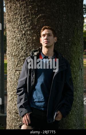 Ritratto in vita di un giovane uomo di etnia latina rossa paraguaiana, all'aperto in giacca blu, al tramonto appoggiato su un albero che guarda la macchina fotografica, stili di vita c Foto Stock