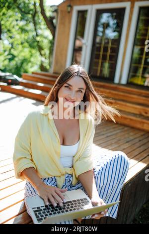 Freelance sorridente seduto con un computer portatile sulla veranda di fronte a casa Foto Stock